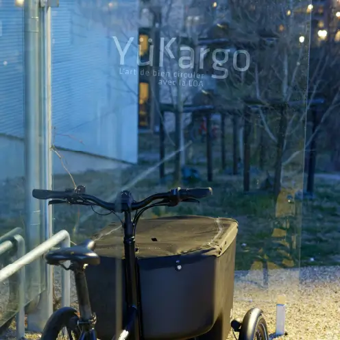 Vélo cargo noir Yükargo avec caisson avant garé derrière une baie vitrée donnant sur un jardin, illustrant l’expérience Yukargo