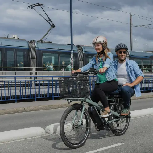 Couple utilisant un vélo cargo électrique vert Yükargo en ville devant un tramway
