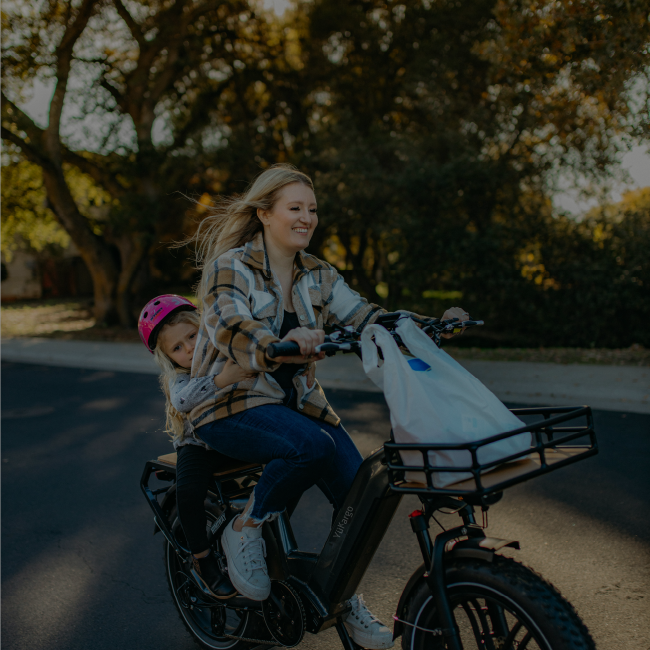 Femme transportant sa fille sur un vélo cargo électrique Yükargo avec sac de courses sur le porte-bagage avant, route avec arbres en arrière-plan