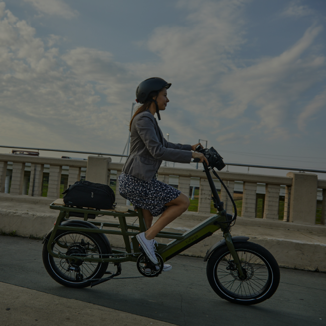 Femme souriante roulant sur un vélo cargo électrique vert Yükargo avec sac sur le porte-bagages arrière, sur un pont