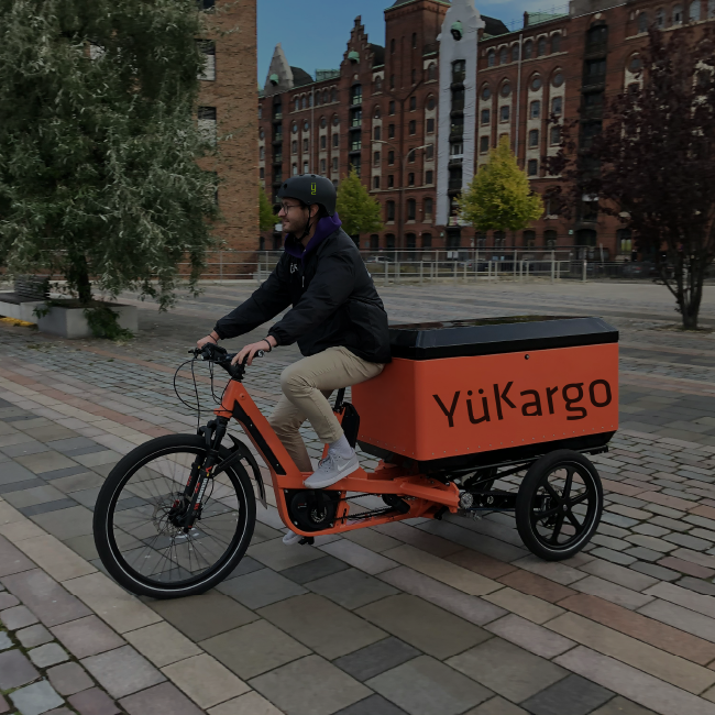 Homme roulant sur un vélo cargo électrique orange Yükargo sur une place urbaine, souriant et détendu